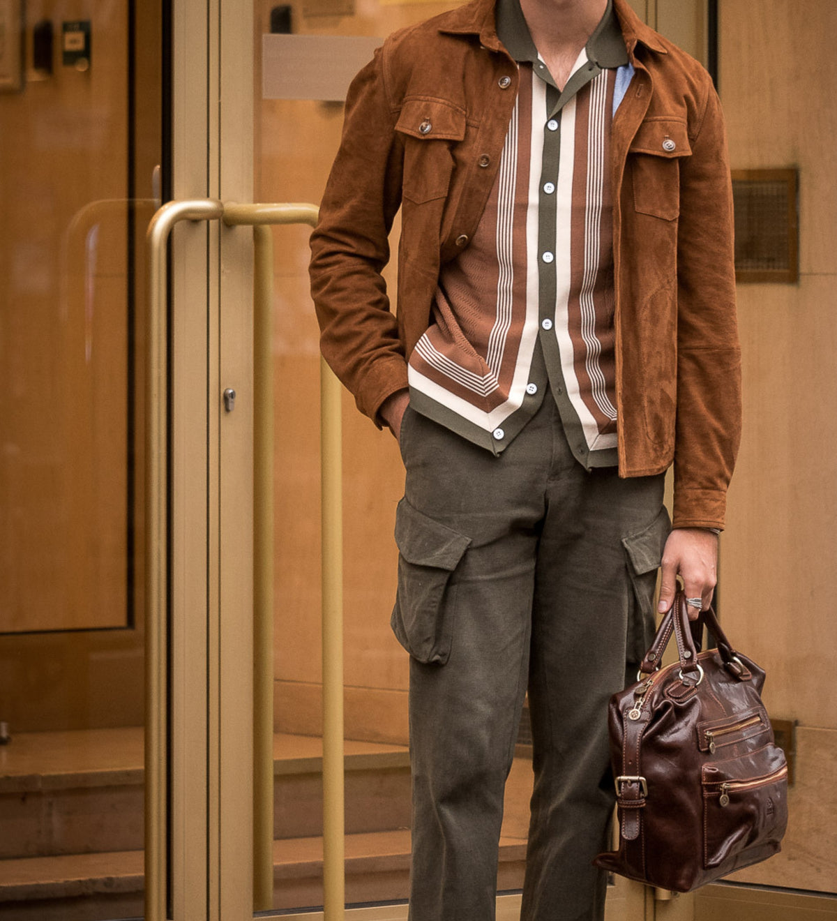 Wearing a suede jacket, a man holds the brown East of Eden leather bag near a doorway